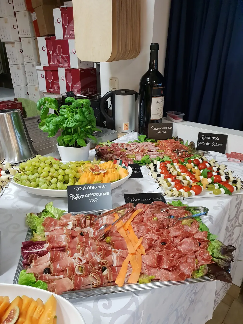 Langes Buffet auf weißen Tischen mit Schinkenplatten, Melone, Trauben und Kreidetafeln