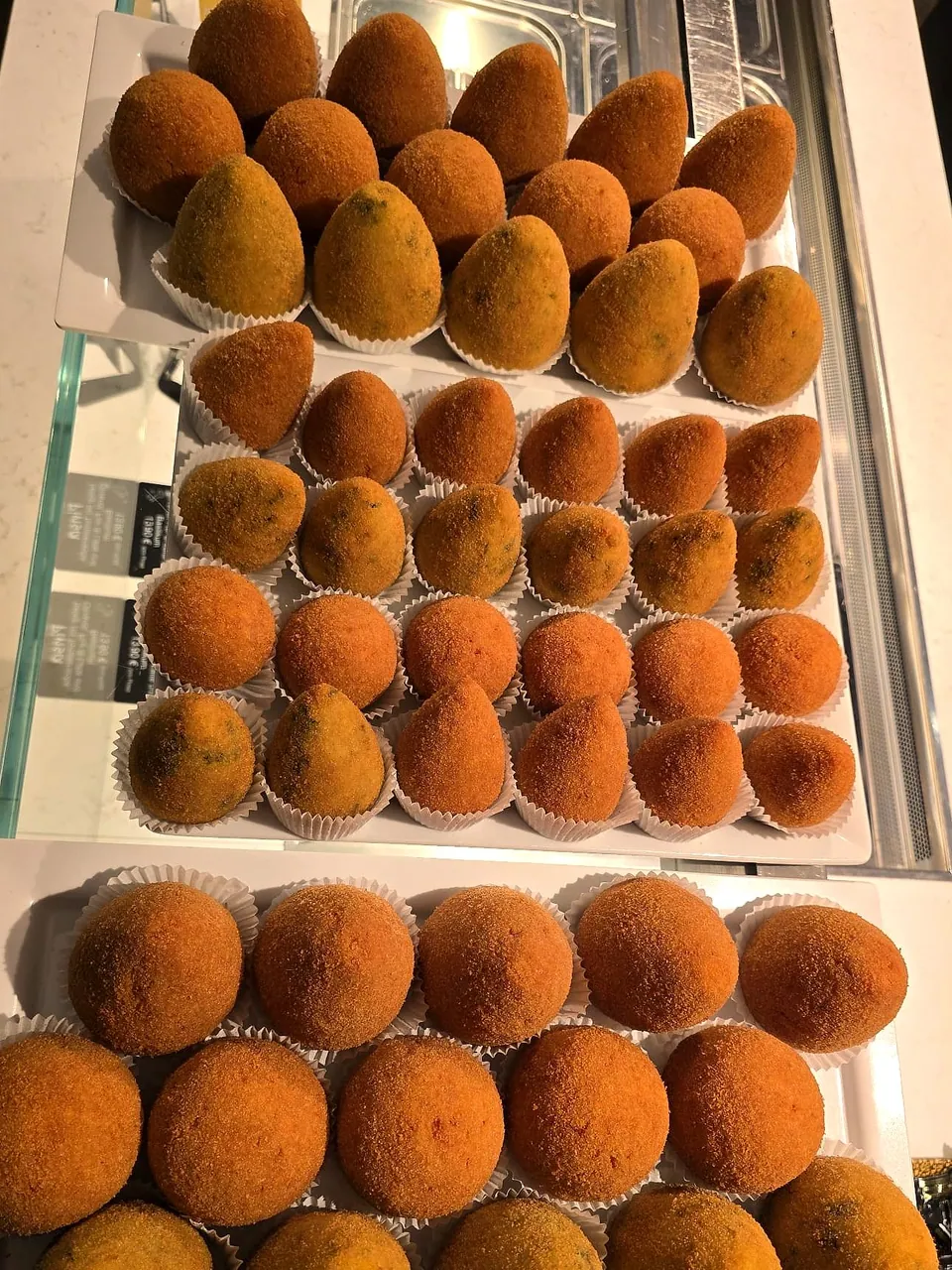 Drei Bleche handgemachte Arancini in der Theke: runde und spitze, goldbraun paniert, einige mit Spinat