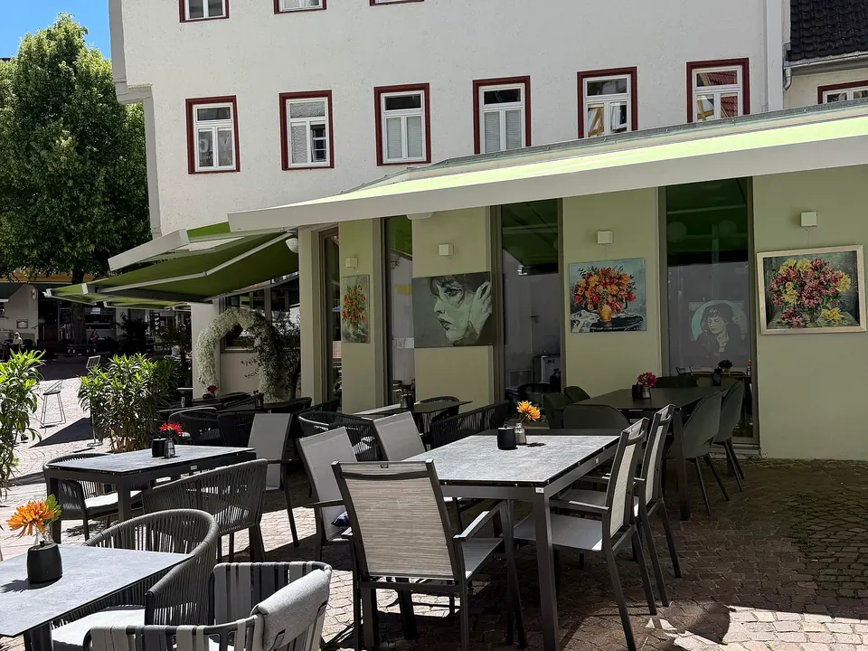 Die Außenterrasse der Markthalle in der Sonne: Tische unter grüner Markise, Gemälde an der Fassade