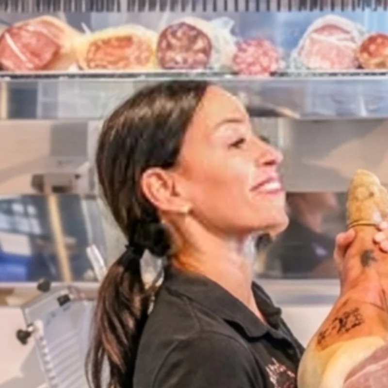 Marisa lacht mit einem ganzen Prosciutto im Arm vor der Wurst-Vitrine